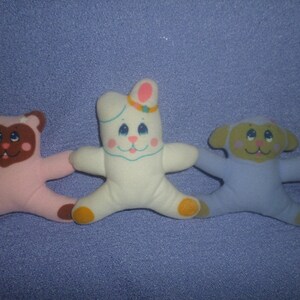 May include: Three plush toys: a pink bear, a white bunny with a pink bow, and a blue lamb. The toys have embroidered faces and are in a playful pose.
