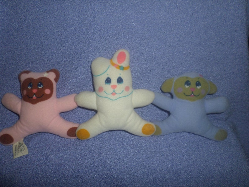 May include: Three plush toys: a pink bear, a white bunny with a pink bow, and a blue lamb. The toys have embroidered faces and are in a playful pose.