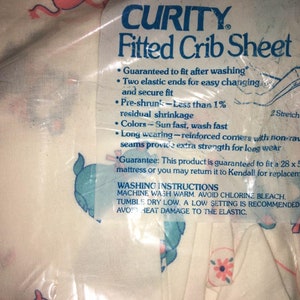 May include: A packaged Curity fitted crib sheet with a pattern of pink and blue elephants. The packaging includes text about the sheet's features, such as being pre-shrunk and sun-fast. The sheet is designed to fit a 28 x 52 inch mattress.