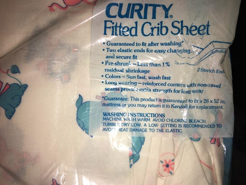 May include: A packaged Curity fitted crib sheet with a pattern of pink and blue elephants. The packaging includes text about the sheet's features, such as being pre-shrunk and sun-fast. The sheet is designed to fit a 28 x 52 inch mattress.