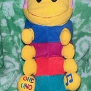 May include: A colorful plush toy caterpillar with a yellow head, blue, pink, and red stripes, and yellow segments. The toy has a musical note on one side and the words "One Uno" on the other.