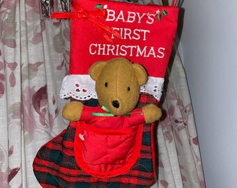 May include: A red Christmas stocking with the text "BABY'S FIRST CHRISTMAS" embroidered in white. The stocking has a plaid pattern and a small teddy bear attached. A red bow and white lace trim the top.