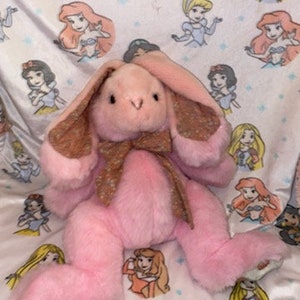 Peut inclure: Un lapin en peluche rose avec des oreilles brunes et dorées et un nœud papillon brun et doré. Le lapin est assis sur une couverture blanche avec un motif de princesses Disney.