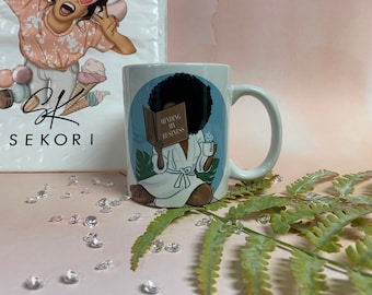 May include: White ceramic mug with a black afro woman illustration reading a book that says "Minding My Business" and holding a cup of tea. The mug has a white handle and is set against a light blue background. A shopping bag with the word "SEKORI" is in the background.