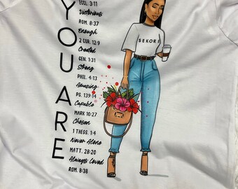 May include: White t-shirt with the words "YOU ARE" in large black letters on the left side. A graphic of a woman in a white shirt with "SEKORI" printed on it, blue jeans, and holding a purse with flowers is on the right.