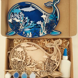 May include: A layered wooden whale art kit with a blue and white ocean scene. The kit includes a wooden whale cutout, paint, glue, and a paintbrush, all inside a cardboard box. The finished whale art features fish, coral, and a starfish.