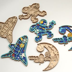 May include: A collection of layered wooden space-themed cutouts. The set includes two astronaut figures, two space shuttles, and two crescent moon designs with planets and satellites. The cutouts feature blue, tan, and white colors, with intricate details.