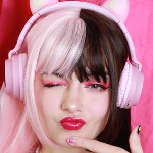 May include: A person with a two-toned wig, half pink and half dark brown, wearing pink headphones with cat ears. The person is winking and wearing pink makeup and lipstick. The background is a pink curtain.
