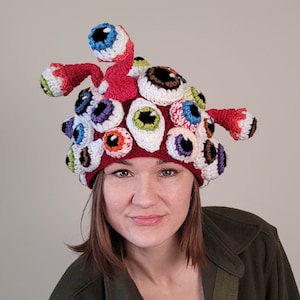 Puede incluir: Un gorro de punto rojo con una variedad de ojos coloridos. El gorro está hecho de hilo y tiene un diseño único y caprichoso.
