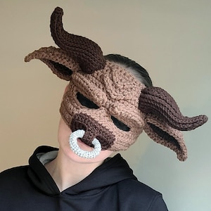 Puede incluir: Una máscara de toro de crochet marrón y negro con un anillo de nariz plateado. La máscara la lleva puesta una persona que lleva una sudadera con capucha negra.