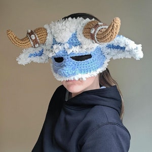 Puede incluir: Una máscara de crochet azul y blanca con cuernos marrones. La máscara la lleva puesta una persona con el pelo oscuro y una sudadera con capucha negra.
