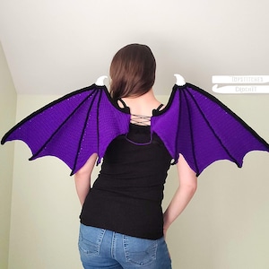May include: Crocheted purple and black bat wings with white tips. The wings are attached to a black top with a lace-up back. The wings are made of a soft, textured yarn and are designed to be worn as a costume accessory.