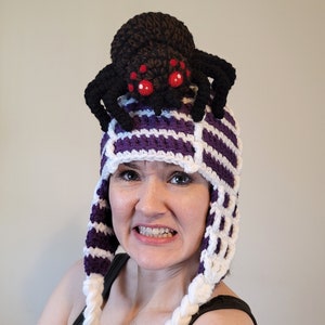 Puede incluir: Un gorro de punto de rayas moradas y blancas con una araña de punto negro en la parte superior. La araña tiene ojos rojos.