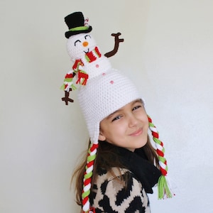 Op de afbeelding: Een witte gehaakte muts met een sneeuwpop erop. De sneeuwpop heeft een zwarte hoge hoed, een rood-groene sjaal en een wit gezicht met zwarte ogen en een rode glimlach. De muts heeft lange rode, witte en groene vlechten.