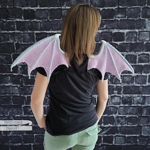 Puede incluir: Alas de murciélago de ganchillo en rosa y gris, usadas sobre los hombros. Las alas tienen un patrón detallado y texturizado y están sujetas con correas negras. La imagen incluye el texto "Topstitches Crochet".