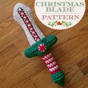 Puede incluir: Un juguete de espada de Navidad de crochet con una hoja blanca, un mango verde y rayas de caramelo rojo y blanco. La espada está sobre una superficie de madera.