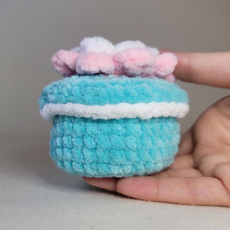 May include: A small, crocheted box with a light blue body, white trim, and a pink and white flower on top.