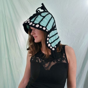 May include: A handmade crochet butterfly hood in black, light blue, and white. The hood features large, wing-shaped panels in light blue with black outlines and white accents. The hood is designed to be worn on the head.