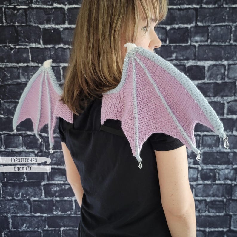 K&ouml;nnte beinhalten: Geh&auml;kelte Drachenfl&uuml;gel in zartem Rosa und Grau. Die Fl&uuml;gel sind an einem schwarzen Kleidungsst&uuml;ck befestigt und weisen ein zartes, strukturiertes Design auf. Die Spitzen der Fl&uuml;gel sind mit kleinen, durchsichtigen Perlen verziert. Der Text "TOPSTITCHES CROCHET" ist sichtbar.