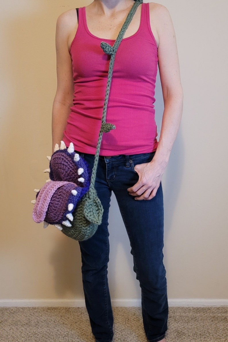 May include: A crocheted green, purple, and pink plant-like bag with a long green strap. The bag is worn over the shoulder of a person wearing a pink tank top and blue jeans.