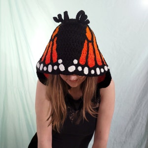 May include: A black crochet hood with orange and white butterfly wing design. The hood features a black base with orange wing patterns and white dots along the edge, resembling a monarch butterfly. The hood has two small antennae-like structures on top.