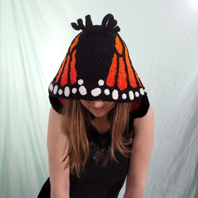 May include: A black crochet hood with orange and white butterfly wing design. The hood features a black base with orange wing patterns and white dots along the edge, resembling a monarch butterfly. The hood has two small antennae-like structures on top.