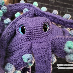 May include: A handmade, purple crochet octopus with large, glittery eyes. The octopus has multiple tentacles adorned with blue and teal pom-poms. The image is a close-up, showcasing the texture of the yarn and the details of the crochet work. The logo "Topstitches Crochet" is in the bottom right corner.
