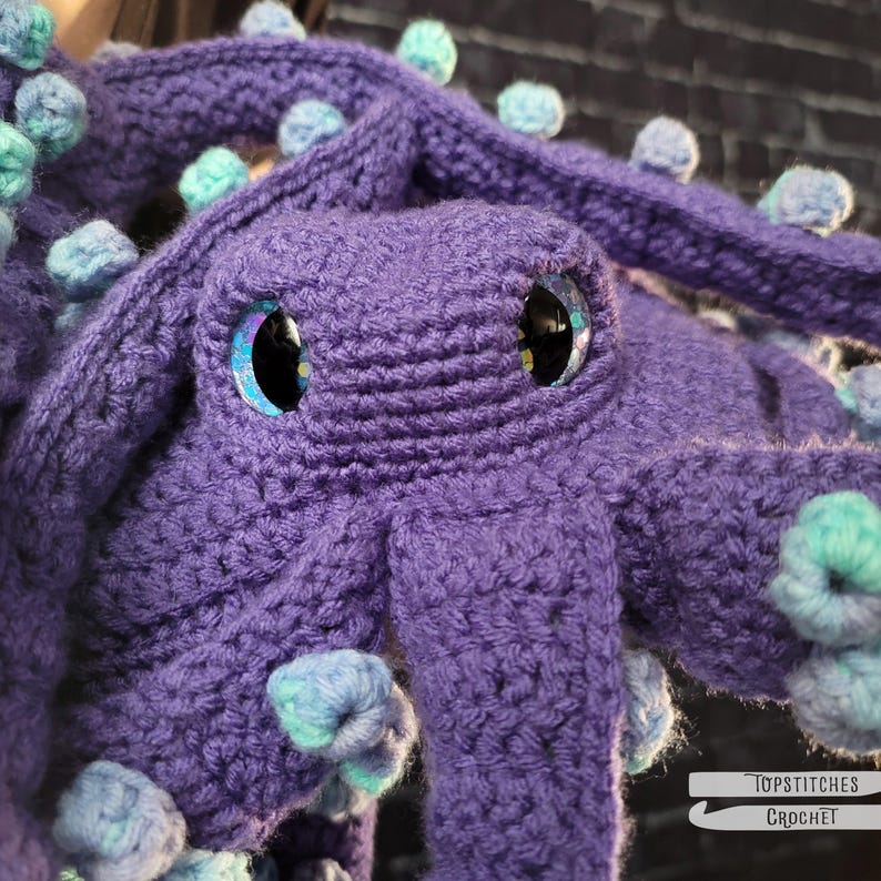 May include: A handmade, purple crochet octopus with large, glittery eyes. The octopus has multiple tentacles adorned with blue and teal pom-poms. The image is a close-up, showcasing the texture of the yarn and the details of the crochet work. The logo "Topstitches Crochet" is in the bottom right corner.