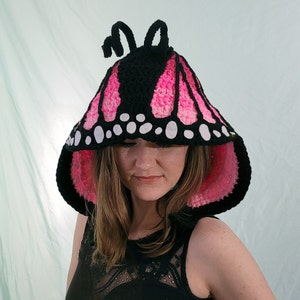 May include: A black, pink, and white crochet butterfly hood. The hood features a black top with pink and white wing patterns, white spots, and two black antennae. The inside of the hood is pink.