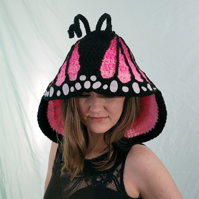 May include: A black, pink, and white crochet butterfly hood. The hood features a black top with pink and white wing patterns, white spots, and two black antennae. The inside of the hood is pink.