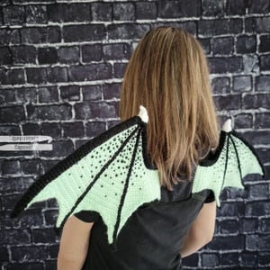 K&ouml;nnte beinhalten: Geh&auml;kelte Drachenfl&uuml;gel in Schwarz und Hellgr&uuml;n, mit schwarzem Rand und kleinen schwarzen Verzierungen. Die Fl&uuml;gel sind an einer wei&szlig;en Basis befestigt. Der Text "Topstitches Crochet" ist auf der linken Seite sichtbar.