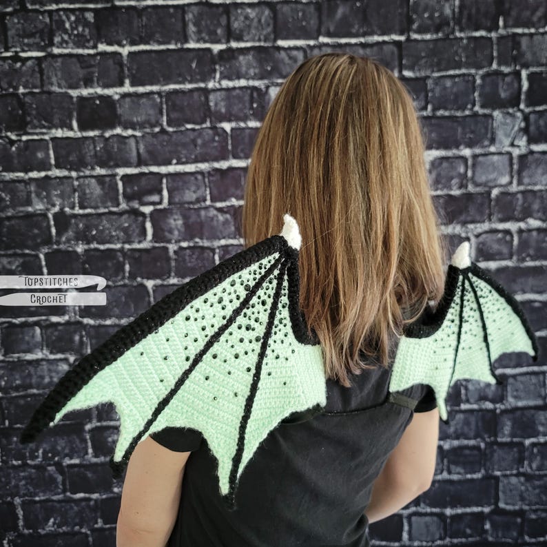 K&ouml;nnte beinhalten: Geh&auml;kelte Drachenfl&uuml;gel in Schwarz und Hellgr&uuml;n, mit schwarzem Rand und kleinen schwarzen Verzierungen. Die Fl&uuml;gel sind an einer wei&szlig;en Basis befestigt. Der Text "Topstitches Crochet" ist auf der linken Seite sichtbar.