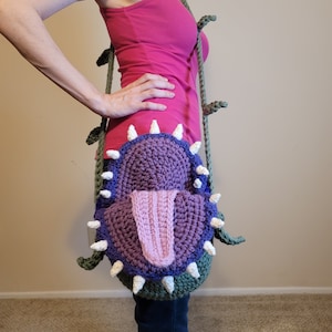 May include: A crocheted shoulder bag shaped like a carnivorous plant with a purple mouth and pink tongue. The bag is green and has a vine-like strap.