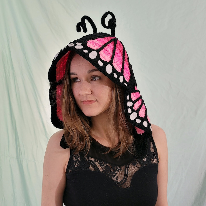 May include: A black and pink crochet butterfly hood with white accents. The hood features a butterfly wing design with pink and black sections, and white dots along the edges. The hood has two black antennae at the top.