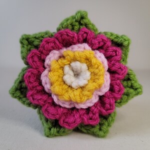 May include: A crocheted flower with green leaves, featuring a pink, yellow and white centre.