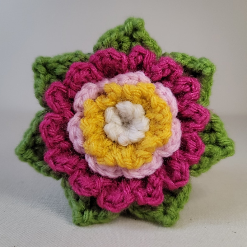 May include: A crocheted flower with green leaves, featuring a pink, yellow and white centre.