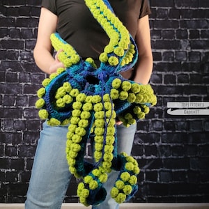 May include: A handmade crochet octopus bag in shades of blue and green. The bag features multiple tentacles with textured bobbles. The bag is held up by a strap. The bag is a unique accessory.