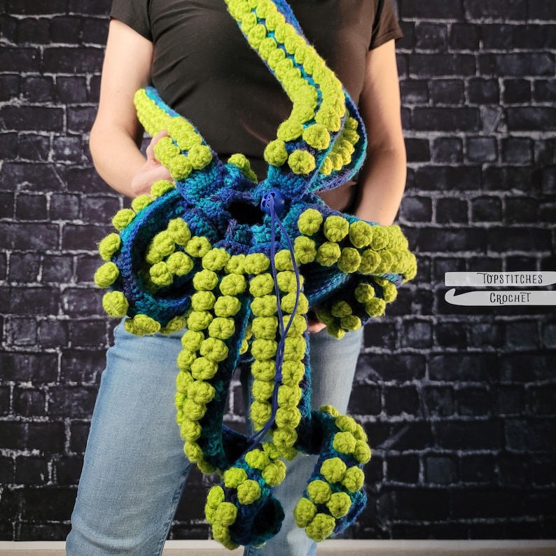 May include: A handmade crochet octopus bag in shades of blue and green. The bag features multiple tentacles with textured bobbles. The bag is held up by a strap. The bag is a unique accessory.