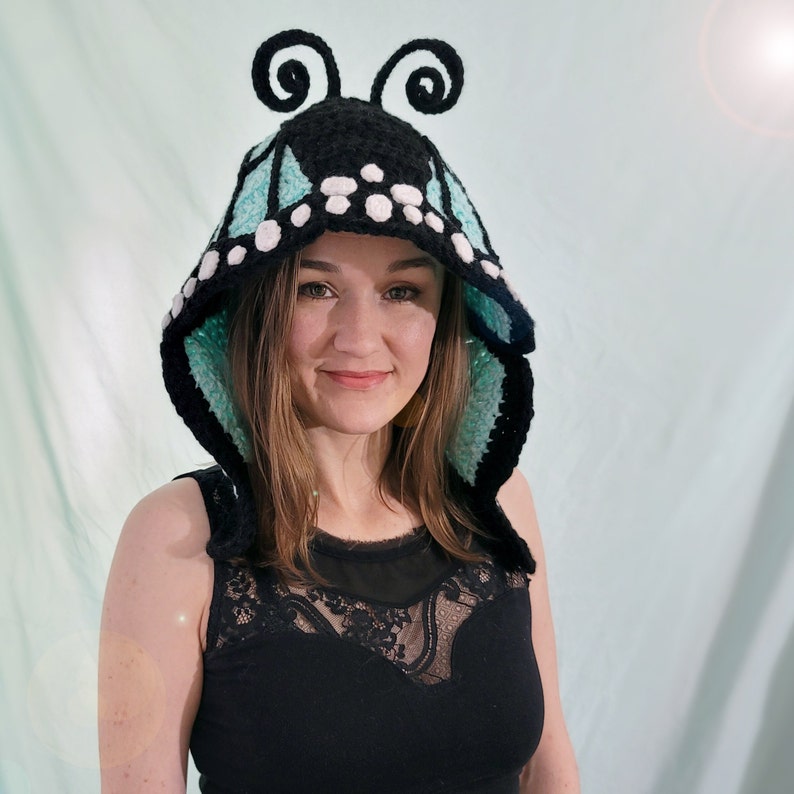 May include: A black and turquoise crochet butterfly hood with white accents and curly antennae. The hood is designed to resemble butterfly wings and is worn on the head. The hood is a unique accessory.