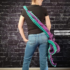May include: A purple and teal crochet octopus shoulder bag. The bag features a long strap with teal bobbles and a large octopus head with teal tentacles. The bag is worn over the shoulder against a black brick wall. The text "TOPSTITCHES CROCHET" is visible.