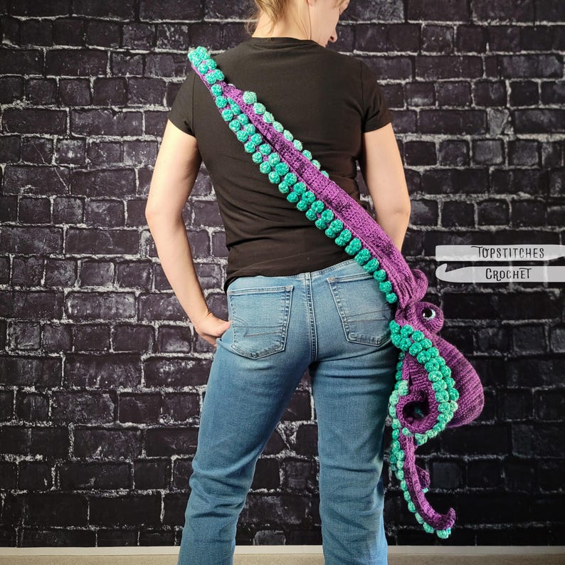 May include: A purple and teal crochet octopus shoulder bag. The bag features a long strap with teal bobbles and a large octopus head with teal tentacles. The bag is worn over the shoulder against a black brick wall. The text "TOPSTITCHES CROCHET" is visible.