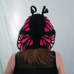 May include: A black and pink crocheted butterfly hat. The hat features a black base with pink wing designs and two black antennae. The hat is worn by a person wearing a black top.