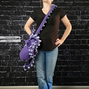 May include: A handmade purple crochet shoulder bag with white accents. The bag features a unique design with a long strap adorned with small, round embellishments. The bag is displayed against a dark brick wall.