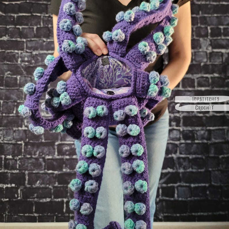 May include: A purple crochet octopus-shaped bag with multiple tentacles. The tentacles are adorned with blue and green bobbles. The bag has a button closure and a patterned interior. The bag is held in front of a brick wall.