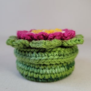 May include: A crocheted flower pot with a pink flower on top. The pot is made of green yarn and has a layered design.