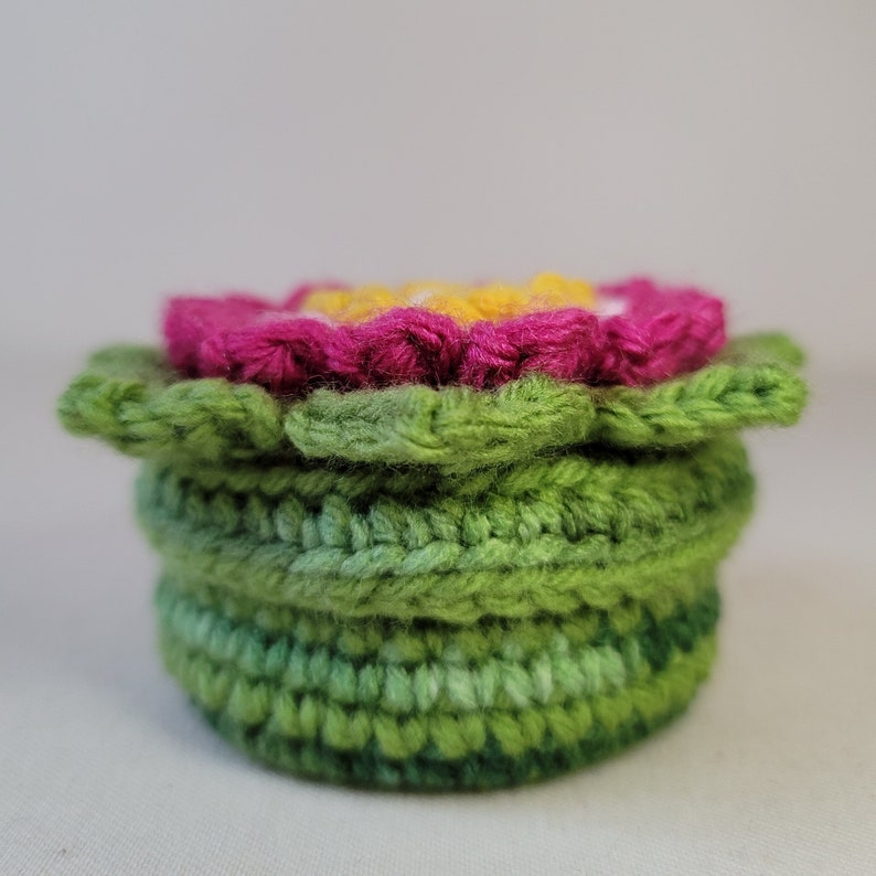 May include: A crocheted flower pot with a pink flower on top. The pot is made of green yarn and has a layered design.