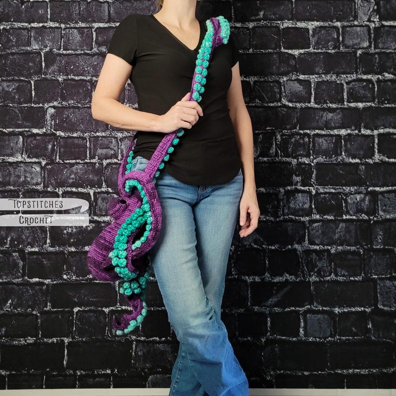 May include: A purple and teal crochet octopus-shaped shoulder bag. The bag features a long strap with teal bobbles and tentacles. The bag is held by a person wearing a black shirt and blue jeans. The bag is made by Topstitches Crochet.