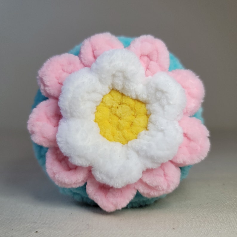 May include: A crocheted flower with pink and white petals and a yellow centre, on a light blue background.
