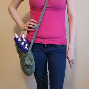 May include: A green crocheted bag shaped like a monster's mouth with purple and white teeth. The bag is worn as a crossbody bag.
