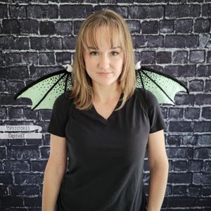 K&ouml;nnte beinhalten: Geh&auml;kelte Drachenfl&uuml;gel in Schwarz, Mintgr&uuml;n und Wei&szlig;. Die Fl&uuml;gel sind an einem schwarzen Geschirr befestigt. Der Text "Topstitches Crochet" ist in der unteren linken Ecke sichtbar. Die Fl&uuml;gel werden auf einer Person vor einer dunklen Backsteinmauer pr&auml;sentiert.